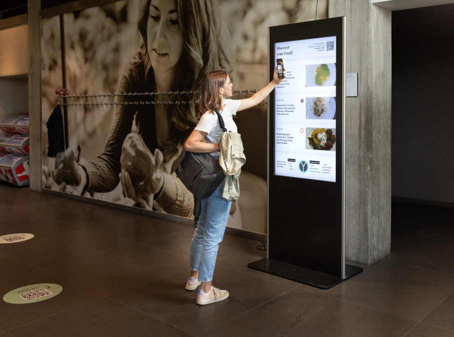 junge Frau scannt mit ihrem Handy einen digitalen Screen mit einem Foodangebot