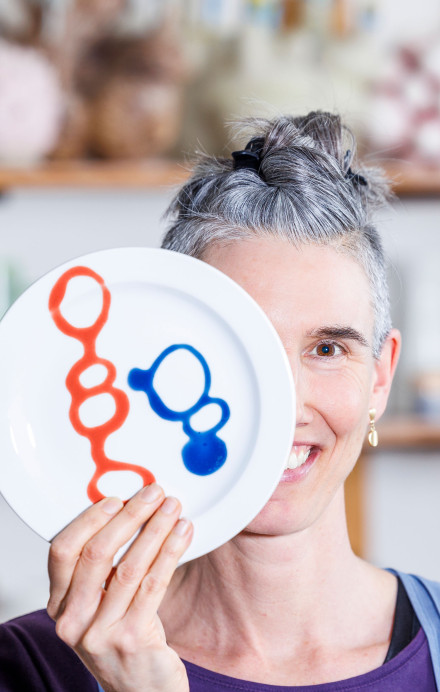 Frau vor einem Regal mit Keramik hält sich einen kunstvoll verzierten Teller vor das Gesicht