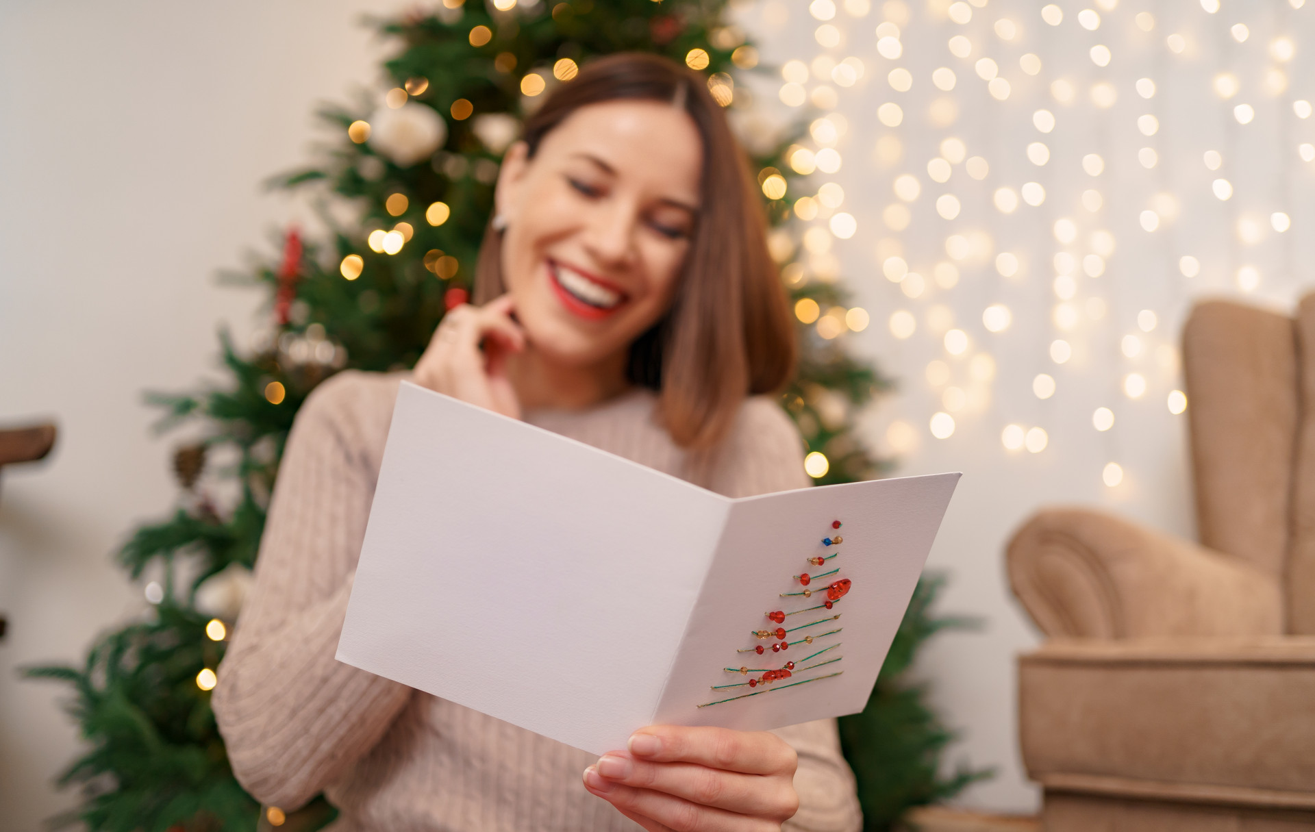 Frau liest Weihnachtskarte und lächelt dabei, hinter ihr ein Weihnachtsbaum und Lichterglanz