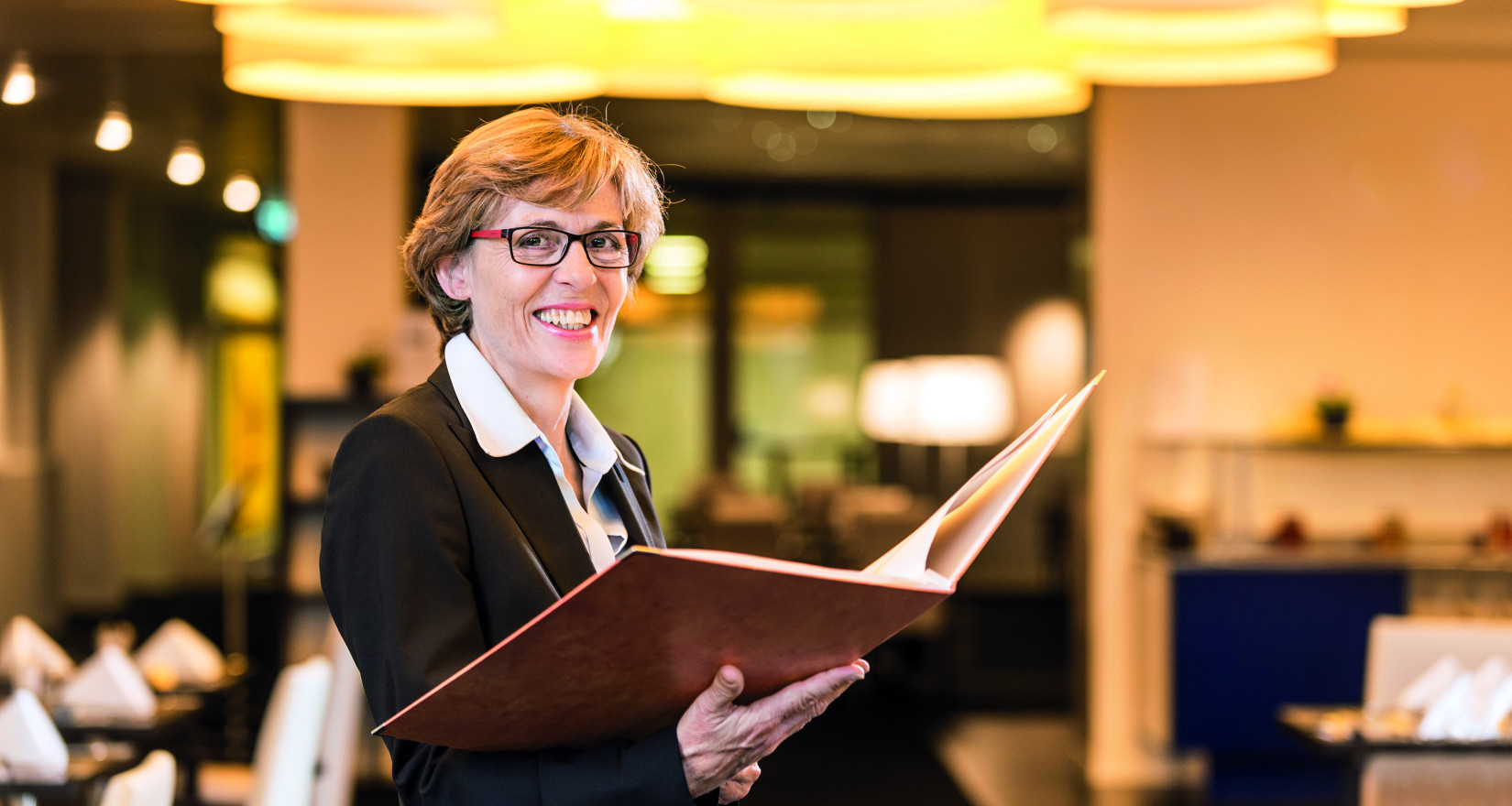 Frau mit einer grossen Speisekarte blickt stehend in die Kamera, hinter ihr ist der Innenraum einres Restaurants zu sehen