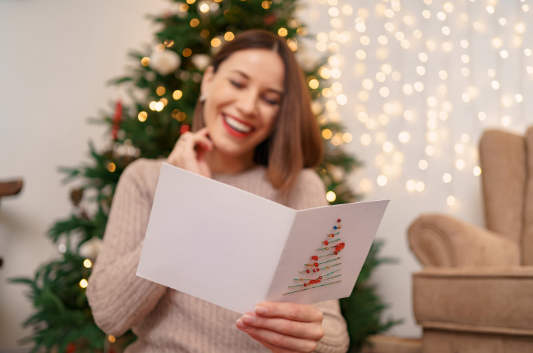 Weihnachtsbaum mit einer jungen, brünetten Frau davor, sie liest begeistert eine Weihnachtskarte