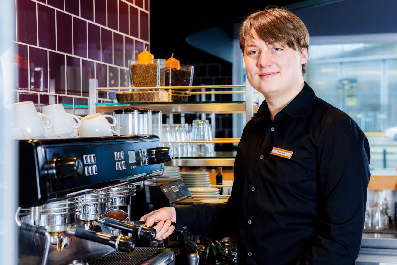 Junger Mann in schwarzem Hemd steht an der Kaffeemaschine und lächelt